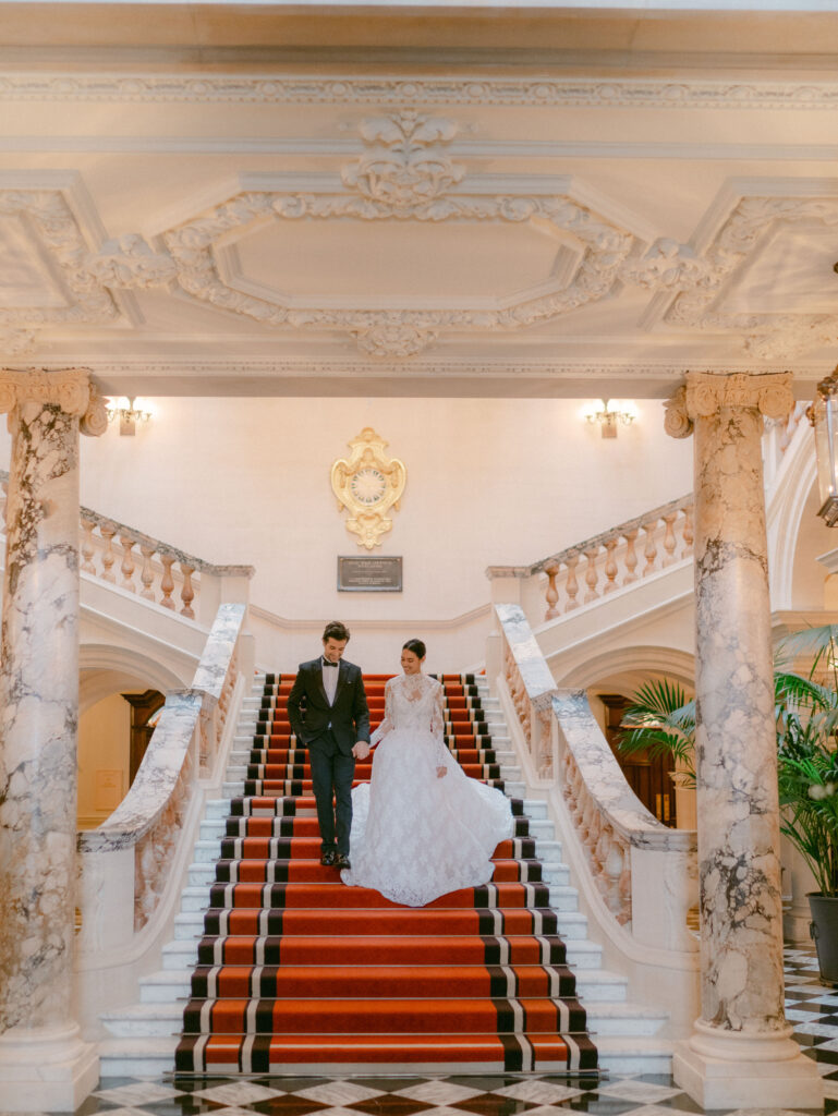 A bride and groom walk down a grand staircase at Raffles London, showcasing the luxury wedding venue's elegant and ornate design.