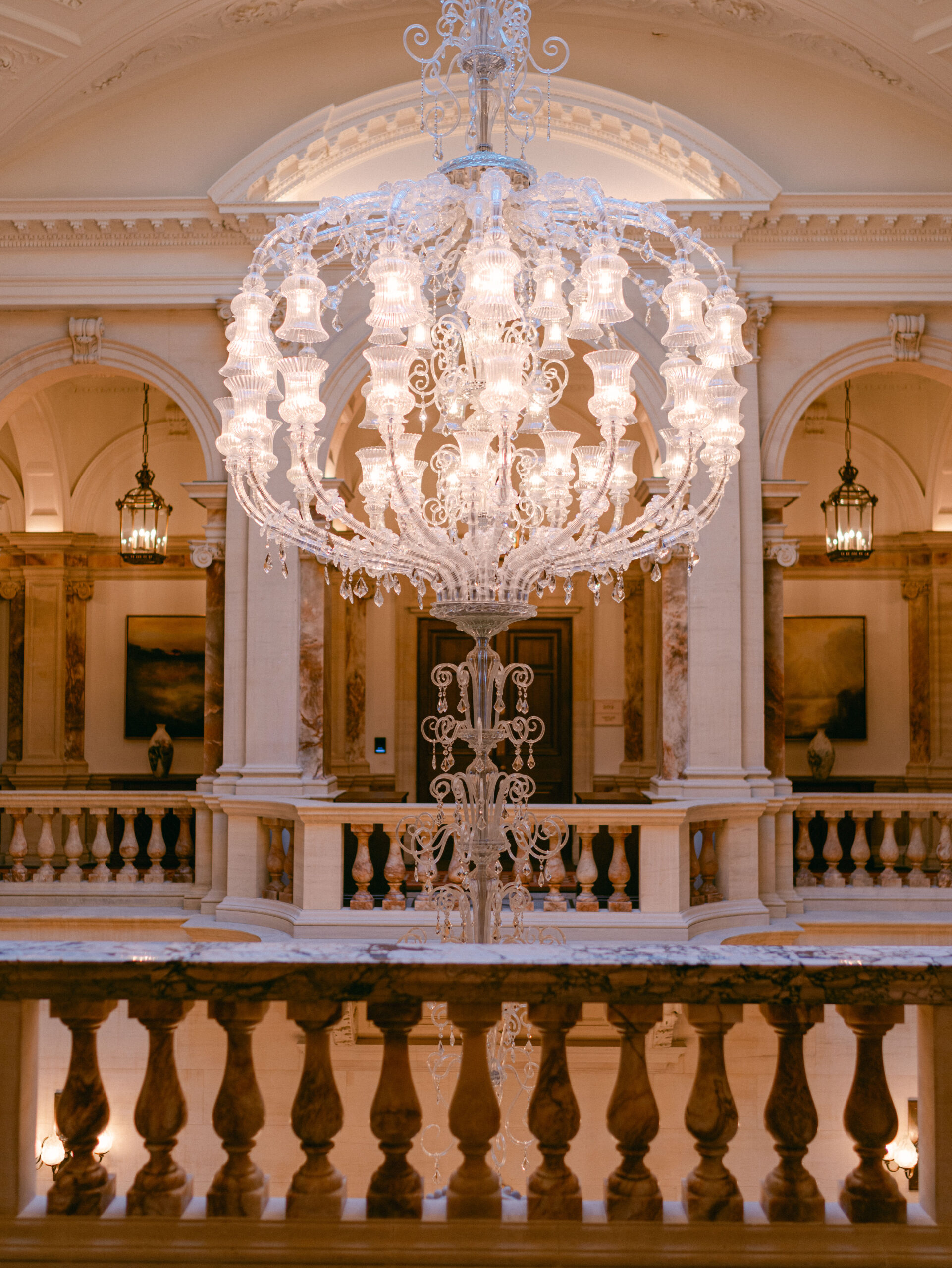 Interior of Raffles London at The OWO Hotel featuring a large opulent chandelier hanging above a grand staircase with intricate detailing, showcasing the venue's luxurious and majestic ambiance.