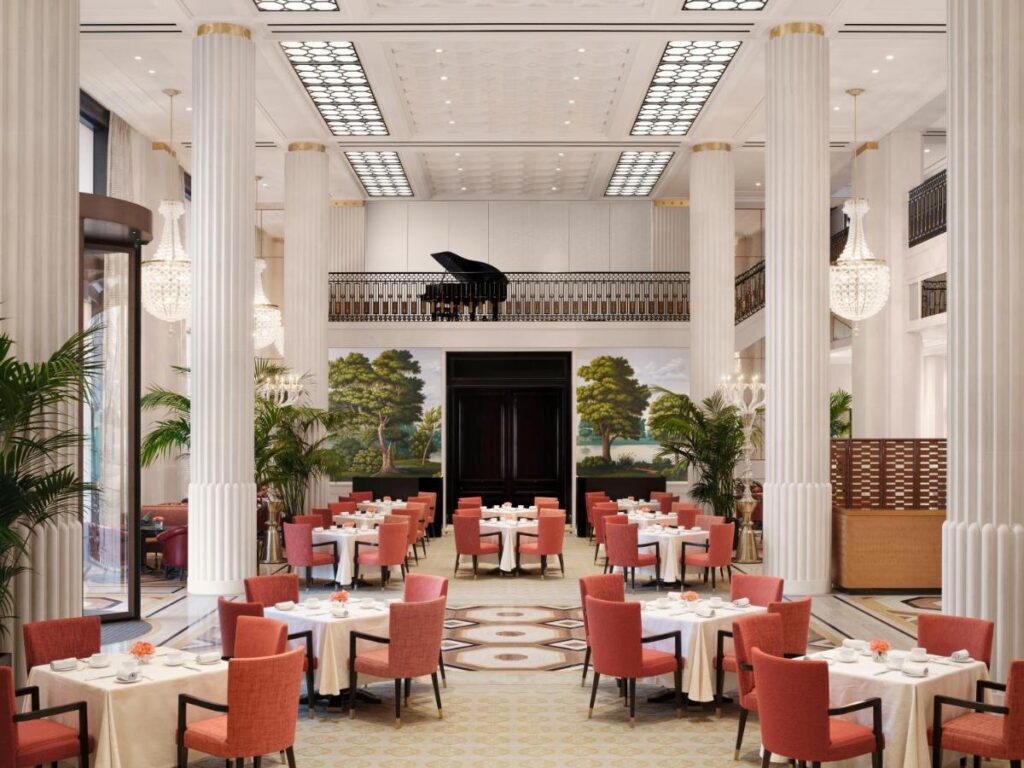 The luxurious dining space at The Peninsula London, featuring elegant columns, a grand piano, and beautifully set tables with red chairs.